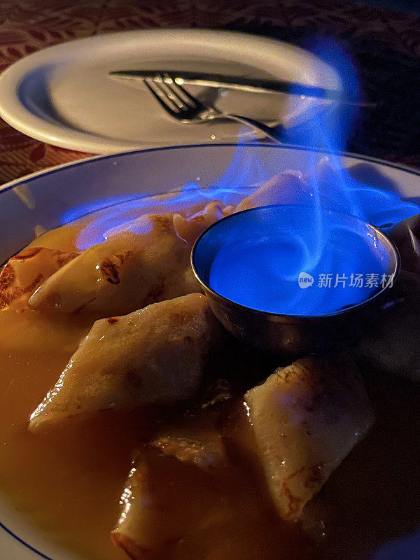 Close-up image of plate of French crêpes (thin pancakes) covered in orange and caramel sauce, Crepe suzette flambe with orange liqueur, blue flame in metal bowl of alcohol, focus on foreground